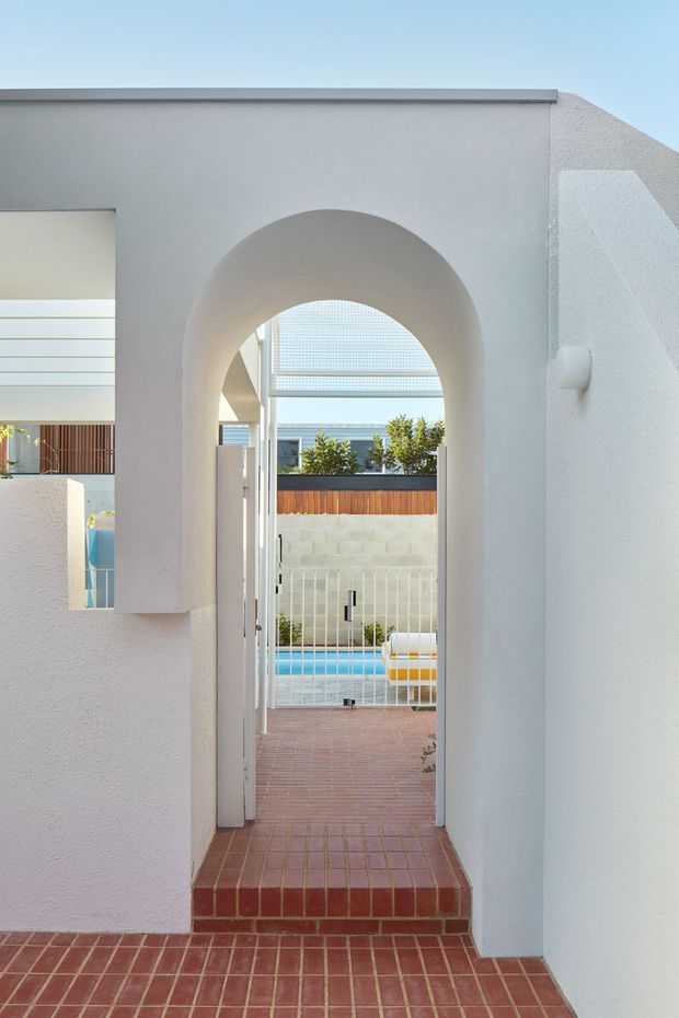 A bright architectural view through a tall rounded archway leading from a red brick-paved area into a small enclosed pool courtyard. White textured stucco walls, a white gate, pale block boundary wall, timber privacy screening, and a turquoise swimming pool are visible beyond. The scene has clean geometric forms with a coastal, resort-like feel.