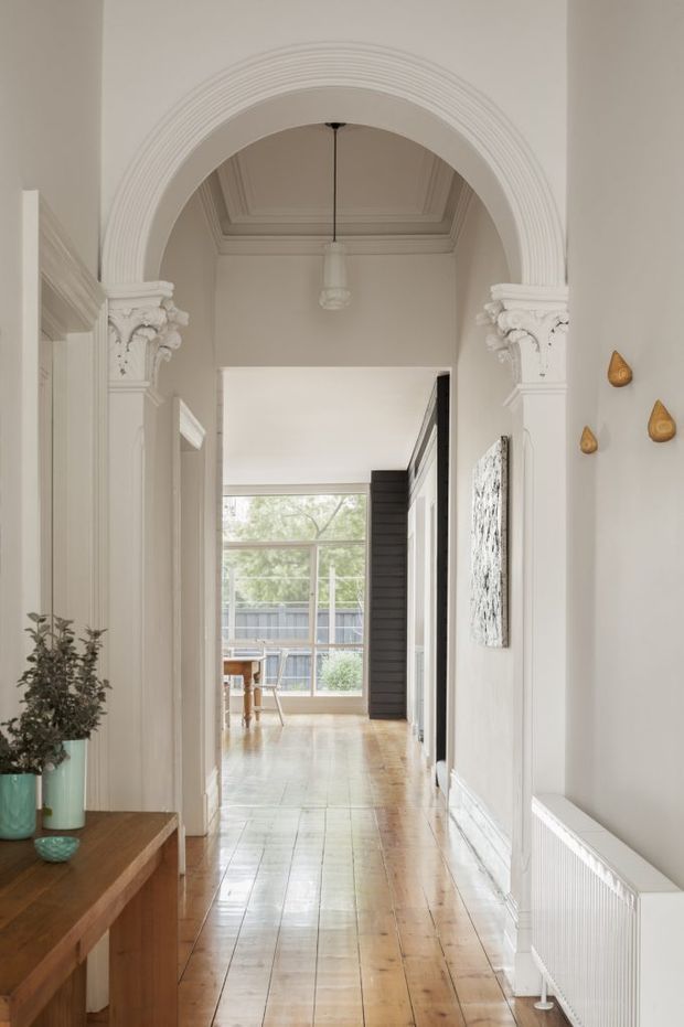 A bright, refined residential hallway featuring a long polished timber floor, white decorative wall mouldings, a large round arch with ornate Corinthian-style capitals, a coffered ceiling, and a pendant light. The corridor leads toward a glazed living or dining area at the rear. A timber console with a potted plant and small decor sits to the left, while a white radiator and subtle wall art line the right side.