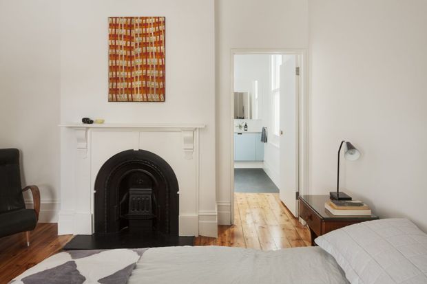 A bright, traditionally detailed bedroom with a white-painted ornate fireplace mantel and black cast-iron fire surround, polished timber floorboards, and a contemporary bed in the foreground. A dark leather armchair sits beside the fireplace, while a simple bedside table and angled task lamp occupy the right side. Through the open doorway, a light-toned bathroom or utility space is visible. The room combines heritage architectural details with restrained modern furnishings and a warm timber-and-white palette.