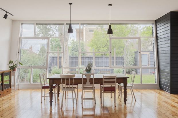 Bright dining area with a long dark timber dining table and mixed wooden chairs, set against extensive floor-to-ceiling-style glazing. The room has polished warm timber flooring, white walls and ceiling, black pendant lights, and a dark horizontal timber feature wall on the right. Large windows overlook a garden and neighbouring residential setting.