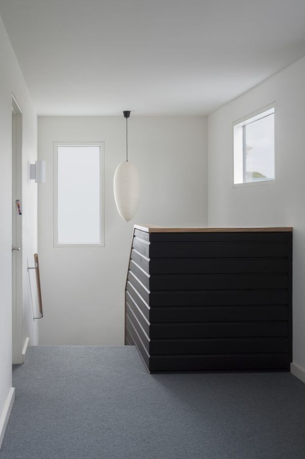 A bright, minimalist hallway or landing with white walls and ceiling, grey carpet flooring, a tall dark built-in or freestanding chest of drawers with a light wood top, and a sculptural white pendant light. A glazed entry door and windows provide soft daylight. The space is clean and sparsely furnished.