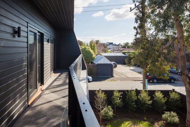 An elevated exterior balcony or walkway along a contemporary dark-clad building, with black horizontal wall cladding, a glazed door, wall-mounted outdoor lights, and a metal balustrade. The view looks over a suburban residential street, driveway, garage roofs, trees, and landscaped planting in daylight.