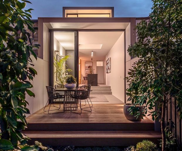A twilight view looking toward a contemporary home's entry and outdoor entertaining terrace. A timber-look deck with black metal dining furniture leads through a large glazed opening into a warmly lit interior foyer, framed by pale rendered or panelled walls and surrounded by dense planting. The facade has a simple rectilinear form with a raised clerestory-like roof element and broad overhanging entry canopy.