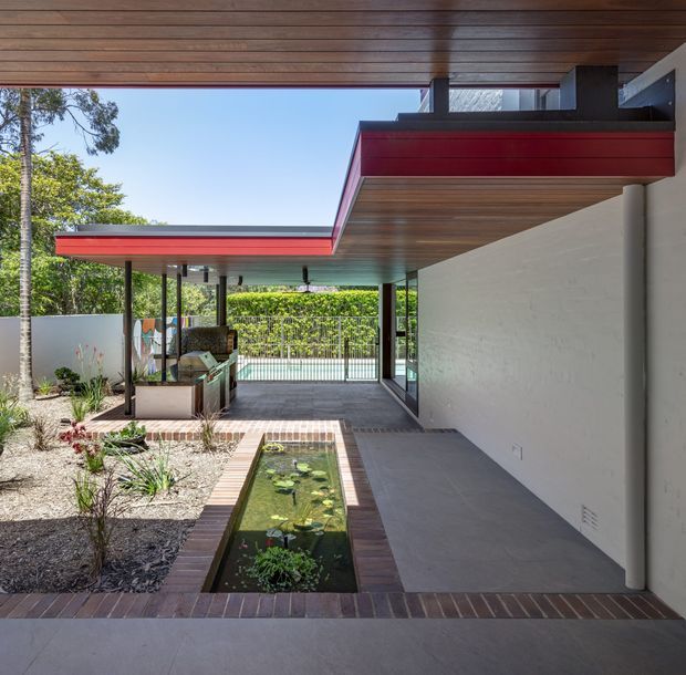 Covered outdoor living and circulation area of a modern home, with a broad timber-lined soffit, flat roof forms edged in red, slender dark posts, white rendered walls, and large grey tiled paving. A narrow rectangular pond with water lilies runs through the foreground, bordered by brick, while landscaped planting and a fenced garden are visible beyond.