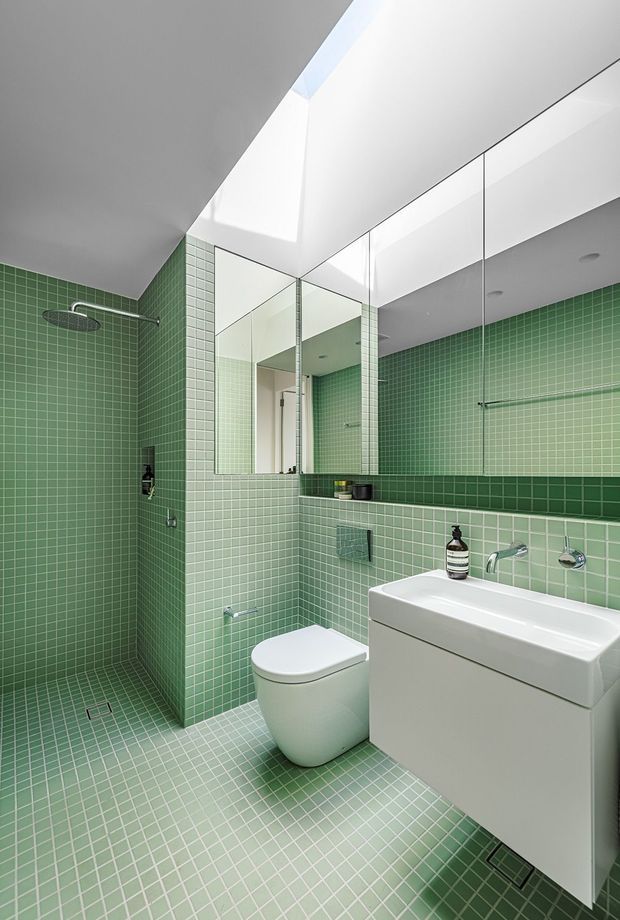 A compact contemporary bathroom with green mosaic tiles covering the floor and walls, including a walk-in shower at left. The room features a wall-hung toilet, a white floating vanity with integrated basin, chrome wall-mounted tapware, a large mirror, and a bright linear skylight or clerestory opening above.