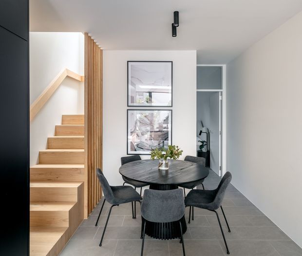 A contemporary dining area beside an open timber staircase. The room features a round dark dining table with four upholstered charcoal chairs, pale walls, large-format grey floor tiles, framed artwork, and black linear ceiling lighting. Vertical timber battens screen the stair, while a doorway opens toward a secondary interior space.