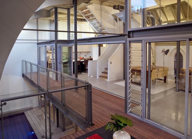 Interior view of a spacious multi-level contemporary home centred on an open stairway and mezzanine walkway. Dark timber decking spans the interior bridges and circulation areas, contrasted with extensive black-framed glazing, metal balustrades, white walls, and exposed structural and roof elements. A kitchen and dining area are visible beyond the glass, with strong visual connections between levels and abundant natural light.