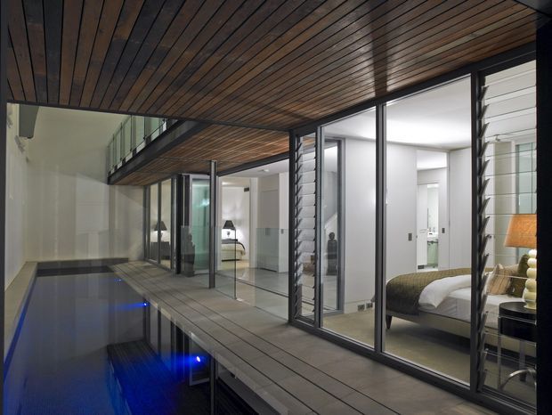 A contemporary interior courtyard or terrace overlooking a dark reflective pool, with a timber-lined soffit and deck walkway. Full-height glass sliding doors open onto bright minimalist interiors, including a bedroom with a bed and bedside lamp. Dark metal window and door frames, white walls, and strong indoor-outdoor connections create a refined, dramatic atmosphere.