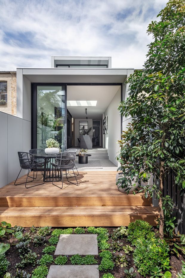 A contemporary urban residential courtyard viewed from a planted garden, featuring a raised timber deck with outdoor dining furniture, steps, and a compact white extension beyond. The extension has large black-framed glazing, a covered entry, a roof-level clerestory or raised volume, and a bright minimalist interior visible through the doors. Dense planting frames the deck and a neighbouring masonry wall is visible at left.