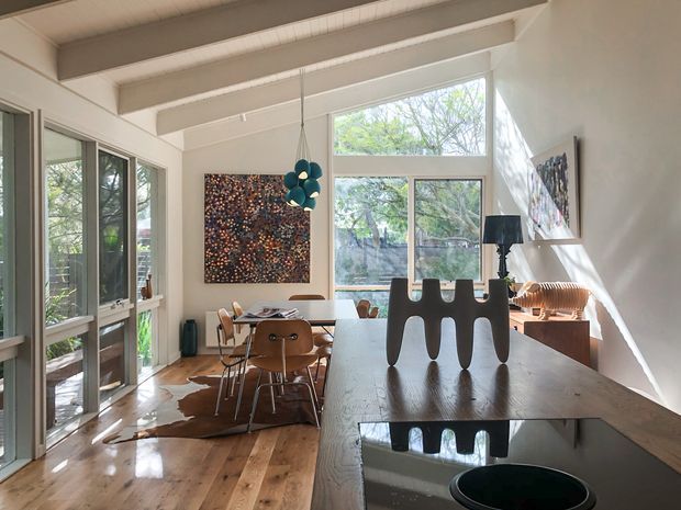 Bright open-plan interior combining a kitchen and dining area, with exposed white-painted ceiling beams, extensive glazing, warm timber flooring, dark timber cabinetry or benchtop surfaces, and mid-century-inspired furniture. A sculptural pendant light, abstract artwork, and strong sunlight create a refined, gallery-like atmosphere.