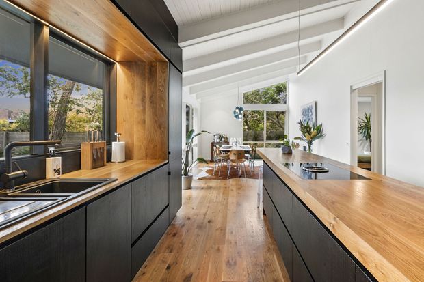 Bright contemporary open-plan kitchen with a long timber island and matching timber benchtop, matte black cabinetry, integrated sink and tapware, and a black-framed window with timber-lined reveal. The space opens into a dining area and is illuminated by large windows, linear pendant lighting, and a white painted exposed-beam ceiling. Warm timber flooring continues through the room.