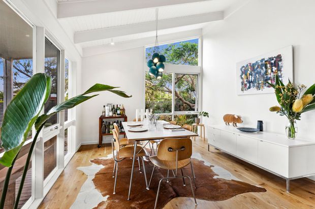 Bright contemporary dining room with a long timber dining table and mid-century style chairs, set on a cowhide rug over warm timber flooring. White built-in cabinetry runs along the right wall beneath abstract artwork and a sculptural floral arrangement. Large glazed doors and windows bring in views of leafy trees, while exposed white ceiling beams and a cluster pendant light add architectural interest.
