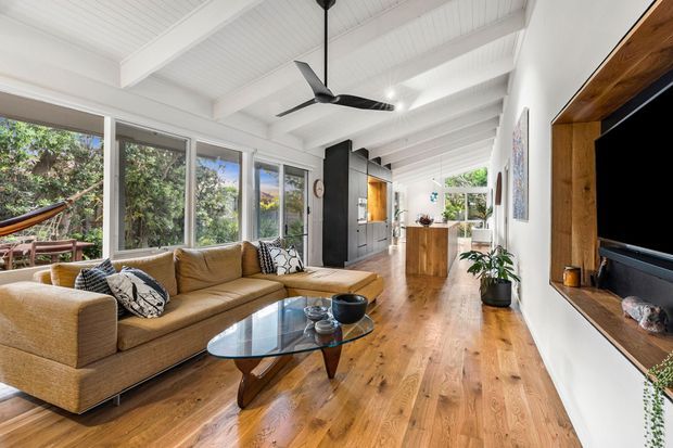 Bright open-plan living room with warm timber flooring, a tan sectional sofa, glass coffee table, exposed white-painted ceiling beams, large windows, a black ceiling fan, and a timber-framed built-in television unit. The space opens toward a kitchen or dining area at the rear and has a relaxed contemporary interior.