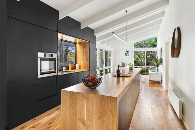 Bright contemporary kitchen with a long natural timber island, matte black full-height cabinetry, integrated appliances, and a timber floor. White exposed ceiling beams and expansive windows and doors bring in abundant daylight and greenery views. The open kitchen extends toward a small dining or living area, with minimalist pendant lighting and restrained decorative accents.