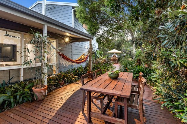 A leafy outdoor timber deck beside a contemporary weatherboard-clad home, furnished with a large wooden dining table and chairs. Potted plants, dense garden planting, a hammock, and a small umbrella-shaded seating area create a relaxed garden entertaining space.