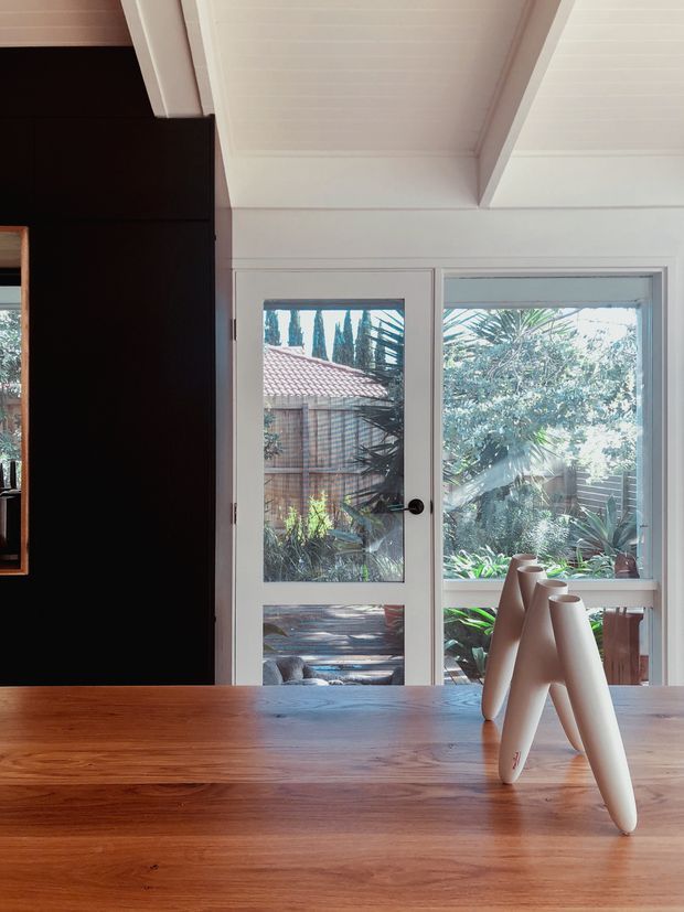 Interior view centered on a warm timber dining table facing white-framed glazed doors and windows opening toward a leafy courtyard or garden. A dark feature wall and partial mirror appear on the left, while the ceiling has white painted timber lining and exposed angled beams. Minimal sculptural decor sits on the table.