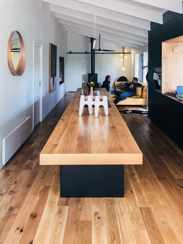 A long, open-plan interior combining a kitchen and living room. A substantial light timber island with a dark base dominates the foreground, matching the wide timber floorboards. Dark cabinetry and a timber-lined open shelving niche frame the kitchen on the right. White-painted exposed ceiling beams, pale walls, a black central fireplace or flue, contemporary pendant lights, and a relaxed seating area create a warm, modern-rustic atmosphere.