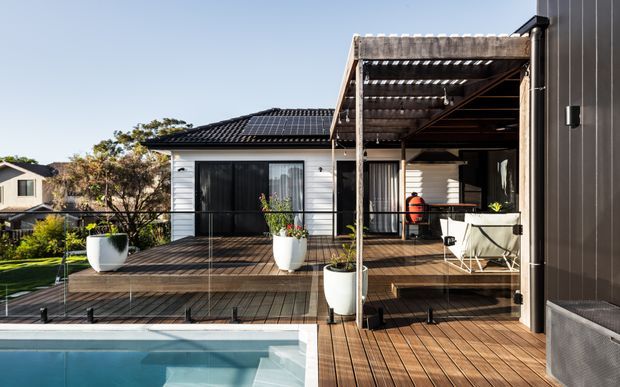 A sunlit outdoor deck and pool area beside a contemporary single-storey home. The expansive timber decking wraps around a swimming pool and is enclosed by frameless glass balustrades. A covered pergola provides an outdoor entertaining area with seating, while the house features dark vertical cladding, white horizontal weatherboards, large glazed openings, and a dark tiled roof.
