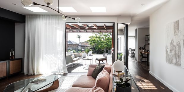Bright contemporary living room with a blush-pink sectional sofa, glass coffee table, dark timber flooring, sheer curtains, and a black feature wall with media cabinetry. Large glazed doors open to a covered outdoor terrace, while skylights and a sculptural linear pendant provide abundant natural and artificial light. A dining or workspace area is visible beyond the living zone.