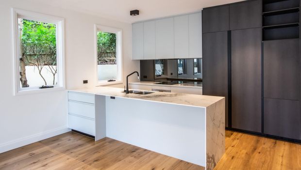 Contemporary kitchen with a large pale stone island, integrated sink and tap, light grey overhead cabinetry, and dark full-height cabinetry with open shelving. Warm timber flooring, white walls, and abundant natural light create a clean, refined contrast between light and dark joinery.