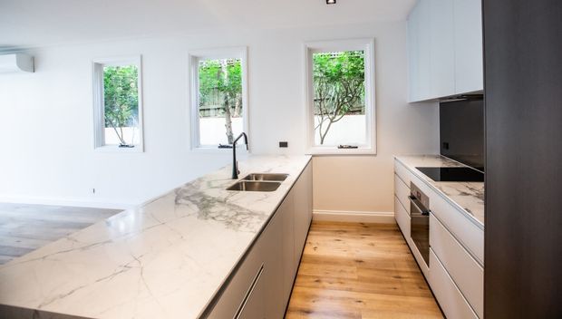 Bright contemporary kitchen with a large white marble-look island incorporating an undermount double sink and slim black tap. The room features white walls, pale timber flooring, white and grey cabinetry, integrated appliances, dark splashback and three narrow windows overlooking greenery.