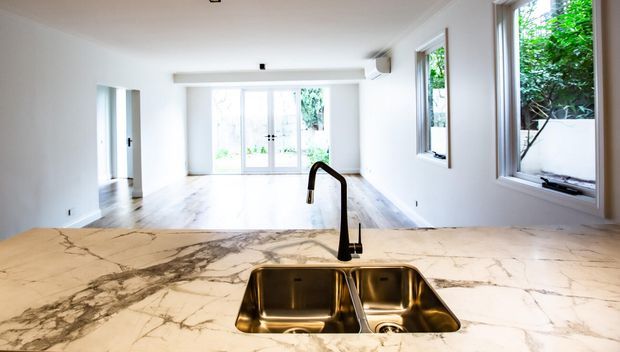 Bright open-plan kitchen and living area viewed from behind a kitchen island. The foreground features a white marble-look stone benchtop with prominent grey veining, an undermount double stainless-steel sink, and a tall black gooseneck tap. The spacious room has pale timber flooring, white walls, large glazed doors and windows, and abundant natural light.