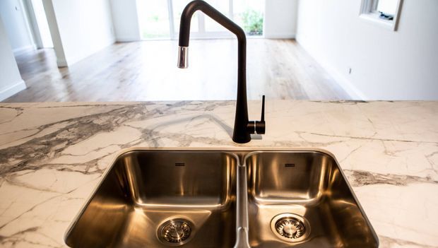 Close-up view of a contemporary kitchen sink and tall gooseneck mixer tap set into a light marble-veined stone benchtop. The double stainless-steel undermount basin dominates the foreground, with pale timber flooring, white walls, and bright glazing visible beyond.
