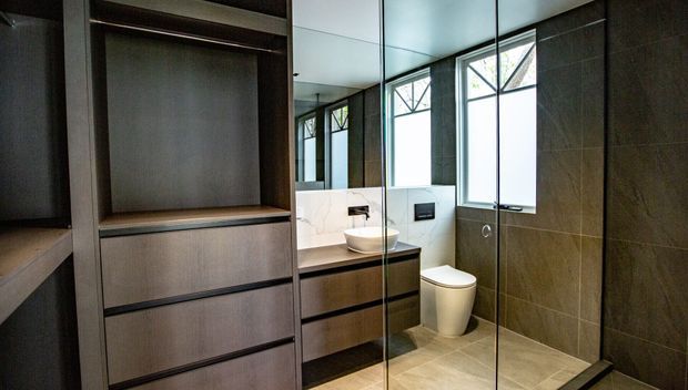 Contemporary bathroom with a walk-in glass shower enclosure, wall-hung vanity with a vessel basin, toilet, mirrored cabinetry, and dark large-format wall tiles. High windows provide natural light, while warm beige floor tiles and dark timber-toned joinery create a refined, modern finish.