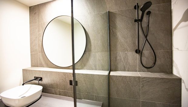 Contemporary bathroom with a wall-mounted white basin, large round mirror, glass shower screen, and black handheld shower fixture. The shower walls and integrated ledge are finished in large-format textured grey tiles, creating a clean, modern look.