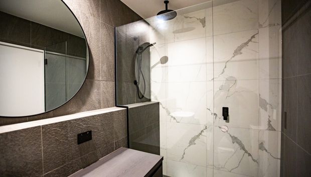 Contemporary bathroom with a walk-in shower, large white marble-look wall tiles with grey veining, darker textured wall tiles, a frameless glass shower screen, matte black fixtures, and a rounded wall mirror above a vanity.
