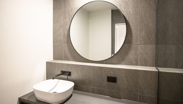 Contemporary bathroom vanity featuring a white vessel basin on a grey stone-look tiled wall, matte black wall-mounted tapware and outlet, a round frameless mirror, and a grey benchtop.