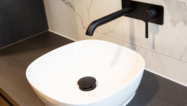 Close-up of a contemporary bathroom vanity featuring a white vessel basin on a dark benchtop, paired with a matte black wall-mounted faucet and black drain. The backsplash is clad in light marble-look tiles with subtle veining, creating a clean, refined contrast with the dark surfaces.
