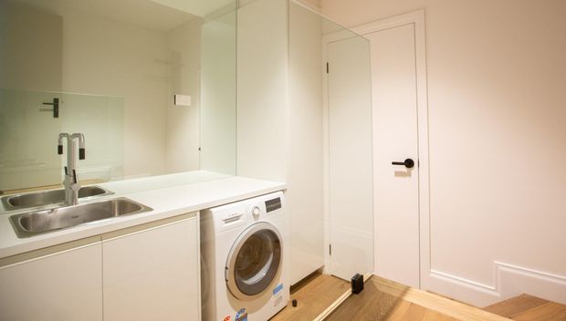 Compact, bright laundry room with white built-in cabinetry and benchtop, stainless-steel double sink, front-loading washing machine, mirrored splashback, and white internal doors. Timber flooring adds warmth to the otherwise neutral fit-out.