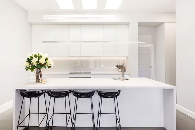 Bright, minimalist white kitchen dominated by a large island with four black wire bar stools. The space features handleless white full-height cabinetry, a white island benchtop, integrated appliances, a pale reflective splashback, recessed task lighting, and a concealed louvred utility door. A floral arrangement provides a small organic accent.