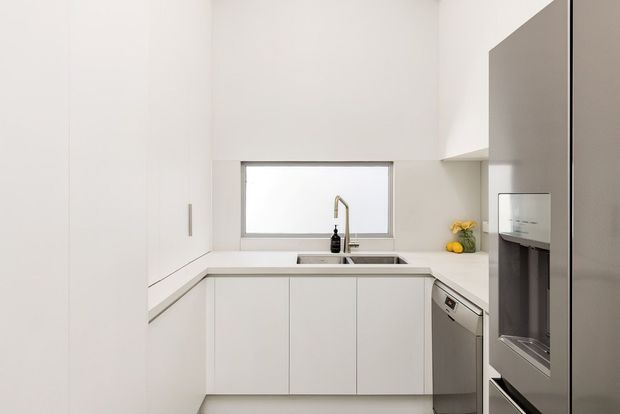 A compact, bright contemporary kitchen with a minimalist white U-shaped layout. Flat-panel white cabinetry runs beneath pale benchtops, with a stainless-steel sink and tap positioned below a horizontal window. A dishwasher sits beside the sink, while a large stainless-steel refrigerator occupies the right side. The space has clean lines, limited decoration, and abundant white surfaces.