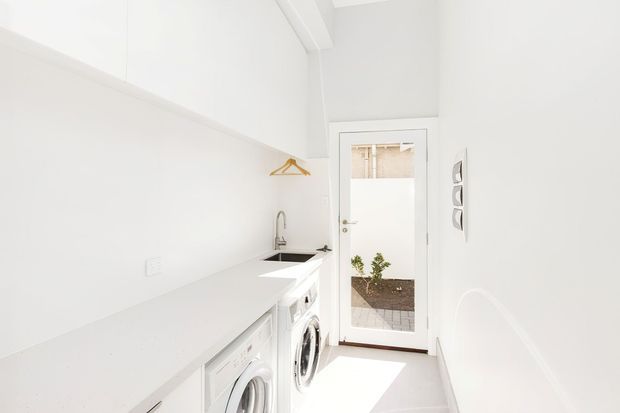 Bright, narrow white laundry room with a long white benchtop, integrated sink and tap, front-loading washing machines, white cabinetry, and a glazed external door opening to a small outdoor area. The room has a clean, minimalist fit-out with abundant natural light.