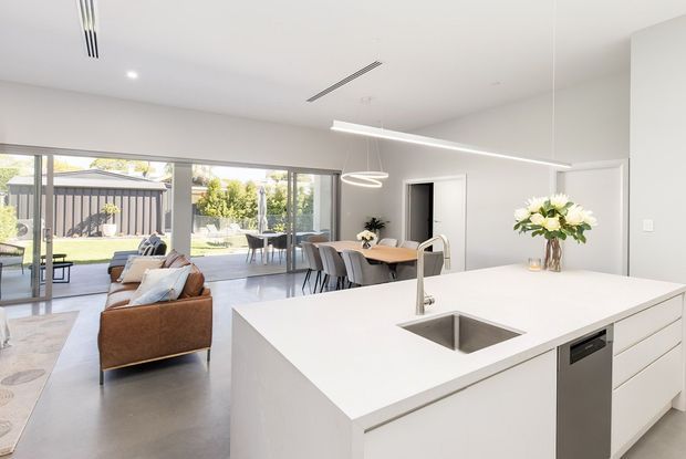Bright open-plan kitchen, dining and living area with a large white island, integrated sink and dishwasher, pale cabinetry, linear pendant lighting, and broad sliding glass doors opening to a landscaped outdoor area. A dining table and upholstered seating are visible beyond the island, creating an airy contemporary family space.