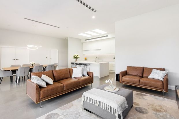 Bright open-plan contemporary living space with a brown leather sofa and matching armchairs arranged around a large upholstered ottoman. A dining area and streamlined white kitchen are visible in the background, with pale flooring, white walls, recessed linear ventilation, skylights, and minimalist pendant lighting.