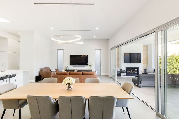Bright, open-plan contemporary interior combining a dining area, living room, kitchen and adjoining outdoor terrace. A large timber dining table with upholstered chairs occupies the foreground, while a neutral-toned living area with sofa, television and fireplace sits beyond. Full-height sliding glass doors open to the terrace, creating strong indoor-outdoor connection. White walls, integrated cabinetry, polished light flooring and minimal suspended lighting give the space an airy, refined appearance.