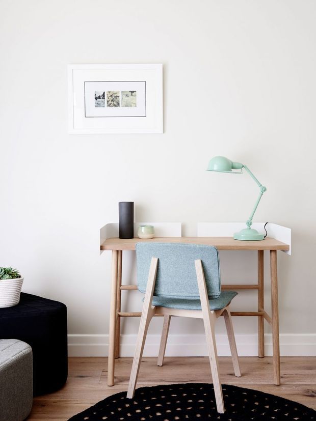A bright, minimalist home office vignette with a pale wood desk against a soft off-white wall. The desk has white side panels, a mint green articulated desk lamp, a dark cylindrical vase, and a small container. A light blue upholstered wooden armchair sits in front, with framed artwork above. Timber flooring, a black round ottoman, a woven plant basket, and part of a grey upholstered seat are visible.