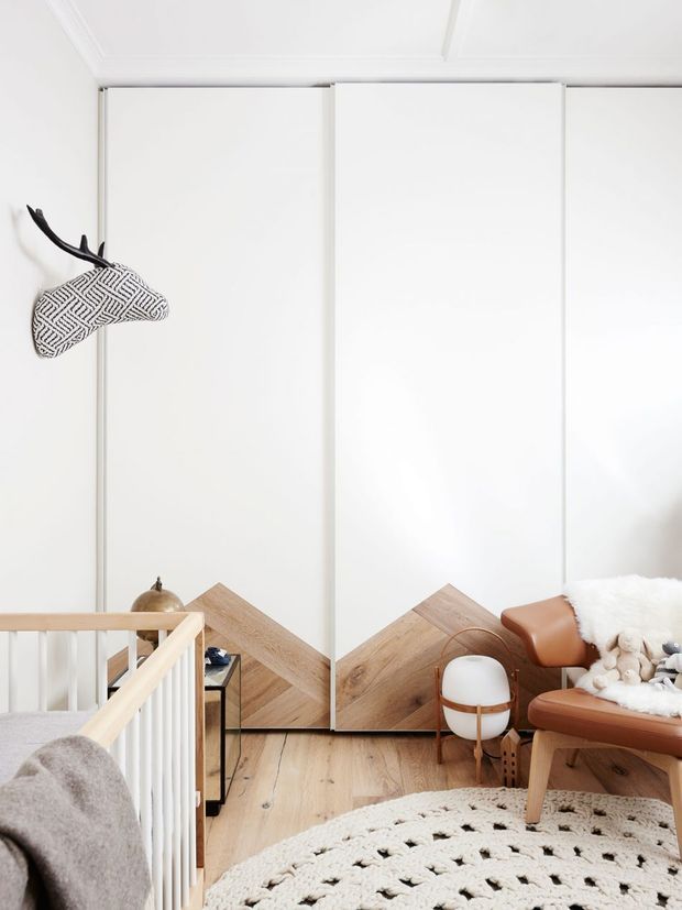 Bright, minimalist upper-level landing or bedroom corner with white wall-to-wall built-in panels, a warm timber floor, and a geometric timber chevron feature at the base of the wall. A white balustrade overlooks the stairwell, while a tan leather lounge chair, small round side table, textured cream rug, soft furnishings, and a sculptural patterned animal wall piece add warmth and character.