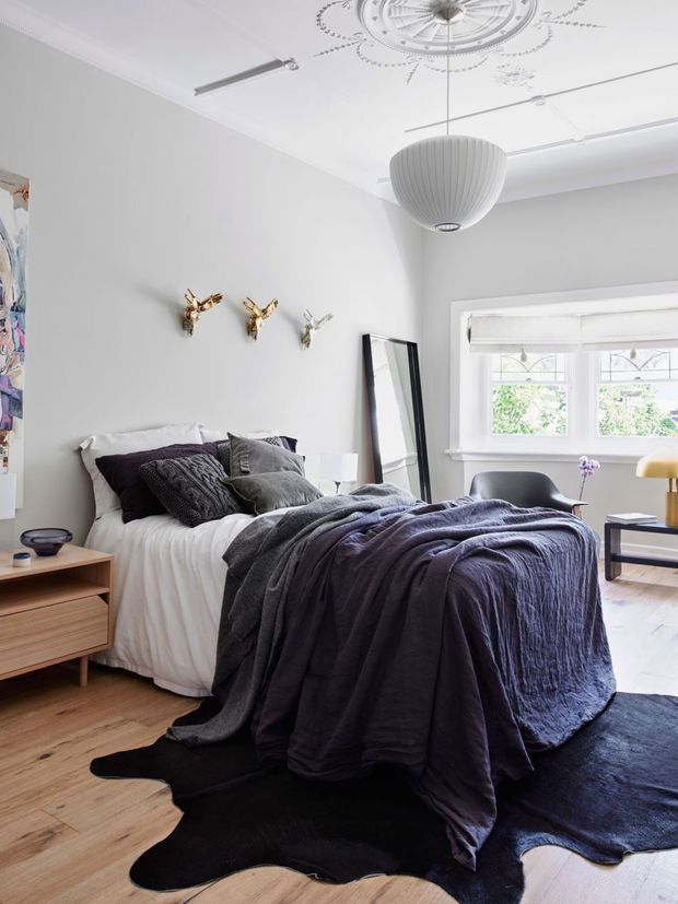 A bright, stylish bedroom with a double bed dressed in layered white, charcoal and deep purple bedding. The room features pale grey walls, a timber floor, a low timber bedside cabinet, a large leaning mirror, a sculptural pendant light, decorative ceiling medallion and cornice detailing, and a broad traditional window with leadlight-style upper panes. A black irregular-shaped rug anchors the bed, while a small seating area is visible near the window.
