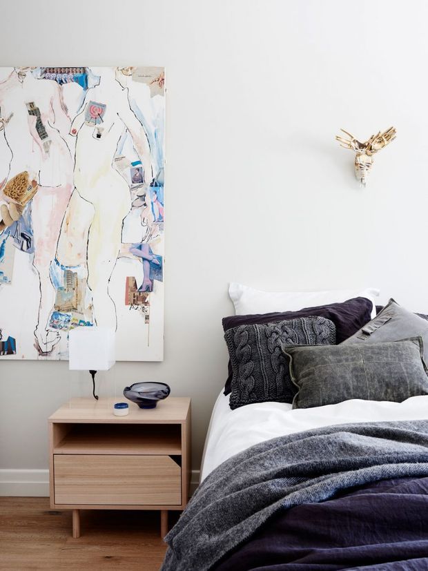 A softly styled bedroom corner with a timber bedside table, table lamp, layered white and charcoal bedding, textured cushions, abstract artwork, and a small sculptural wall light or ornament. The room has a clean, contemporary feel with warm timber flooring and a restrained neutral palette accented by deep purple and charcoal tones.
