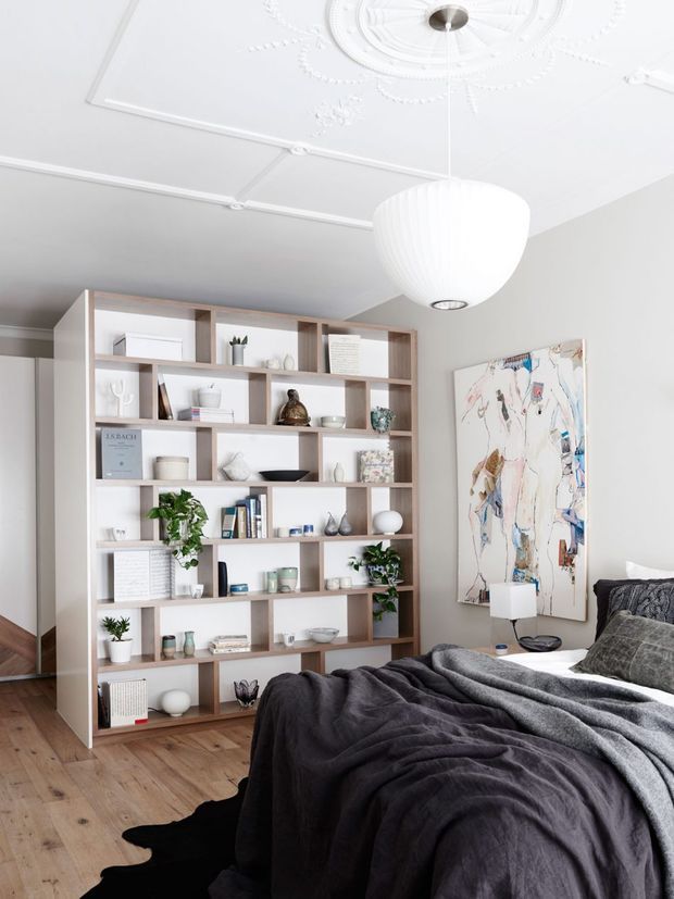 Bright, refined bedroom with a large custom built-in open shelving unit spanning one wall, displaying books, ceramics, artwork and indoor plants. A bed with dark charcoal bedding occupies the foreground, alongside a small bedside table and lamp. The room features pale neutral walls, warm timber flooring, ornate white ceiling detailing with a decorative ceiling rose, and a large white pendant light. A colourful abstract artwork adds contrast.
