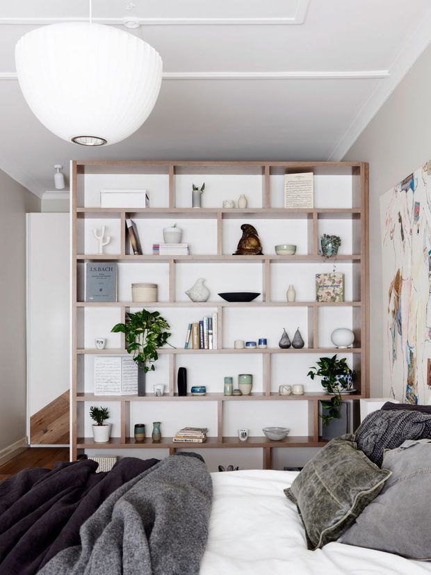 A bright, calm bedroom with a large custom built-in open shelving unit spanning the back wall. The pale timber shelving is styled with books, ceramics, small sculptures, plants, and framed pieces. A large white globe pendant hangs from a decorative white ceiling with moulded trim. The foreground contains a bed dressed in layered charcoal, grey, and white bedding, while a large abstract artwork is partially visible on the right wall. Warm timber flooring and soft neutral walls complete the refined interior.