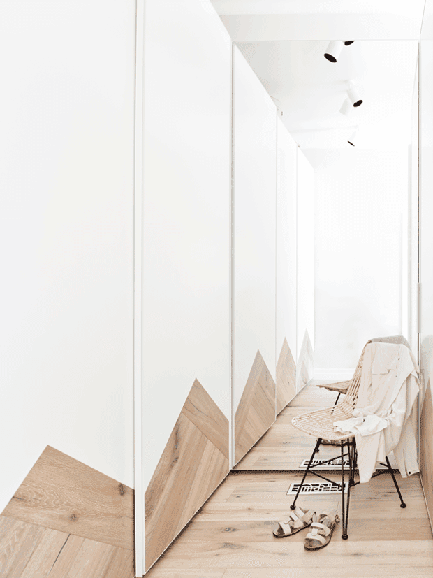 A bright, minimalist white hallway or corridor with full-height mirrored panels creating repeated reflections and a strong sense of depth. The pale timber floor continues along the corridor, with geometric chevron-like timber feature panels at the base of the walls. A woven chair with a draped white textile, sandals, and small black ceiling spotlights add restrained detail. The space is airy and predominantly white with light wood accents.