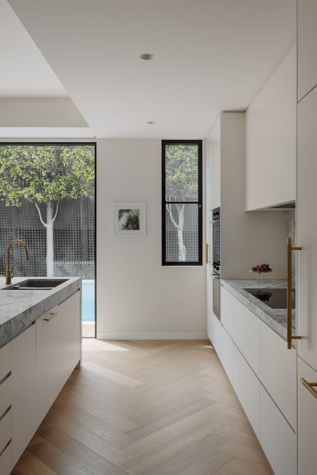 Bright, narrow contemporary kitchen with a central walkway, white full-height cabinetry, pale marble-look stone benchtops, integrated appliances, brass hardware and a pale timber herringbone floor. Large windows and glazed doors bring in views of a leafy courtyard and a glimpse of a pool. The ceiling has recessed downlights and a stepped bulkhead detail.