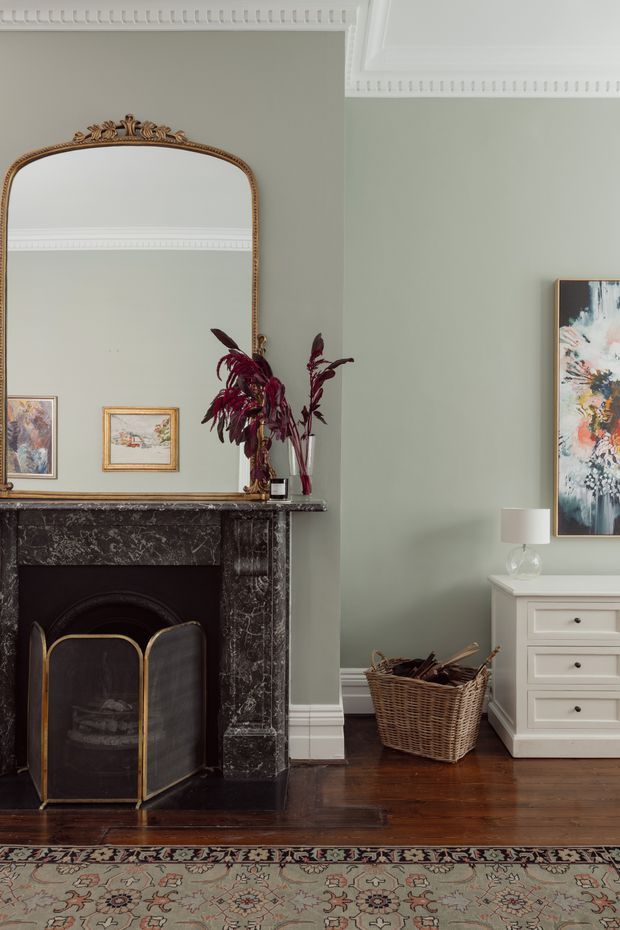 A refined traditional living room with a dark marble fireplace, ornate gilded overmantel mirror, decorative cornices and white trim. The muted sage-green walls contrast with dark timber flooring, a patterned area rug, classic white chest of drawers, woven firewood basket, and restrained antique-inspired furnishings.