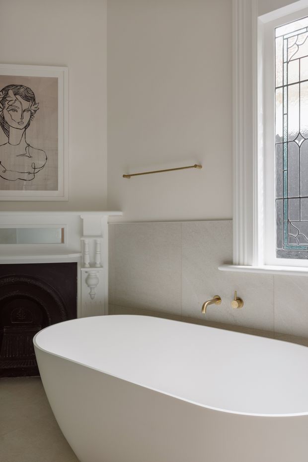 A refined bathroom with a freestanding white oval bathtub positioned beside a traditional dark fireplace. The room has warm greige painted walls, pale stone or porcelain wall tiles, white decorative moulded trim and a tall leaded-glass window. Brushed brass or gold-toned wall-mounted tapware and a slim towel rail add subtle contrast, while a framed monochrome artwork hangs above the fireplace.