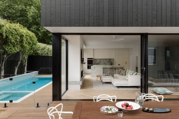 A contemporary rear exterior view of a dark-clad, flat-roofed house opening onto a timber deck and swimming pool. Full-height sliding glass doors reveal a bright open-plan living area with a white sofa, marble-look kitchen island and pale cabinetry. The deck includes an outdoor dining table and white chairs, while established landscaping borders the pool.